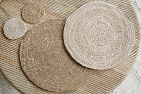 Elevate your minimalist tableware with the Keso Woven Placemats - Brown (Set of 4) by nkuku—handwoven round placemats in varying sizes, beautifully arranged on a light wooden slatted table.