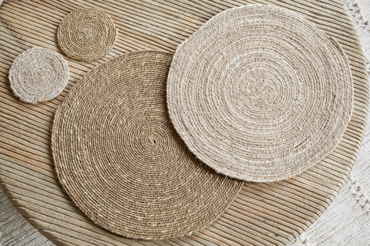 Elevate your minimalist tableware with the Keso Woven Placemats - Brown (Set of 4) by nkuku—handwoven round placemats in varying sizes, beautifully arranged on a light wooden slatted table.