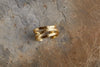 The Katai Ring - Gold by nkuku features three overlapping hammered bands with a gold finish, shown resting on an earth-toned stone beside wispy dry grass.