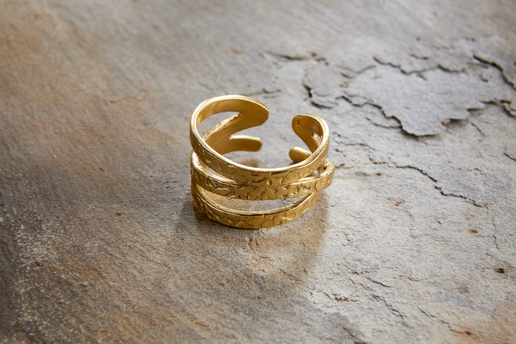 The Katai Ring - Gold by nkuku, featuring a hammered texture and open, layered design, rests on a rugged, stone-like surface.