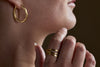 A person touches their chin, wearing the nkuku Katai Ring - Gold and a textured gold hoop earring. The dark background is out of focus.