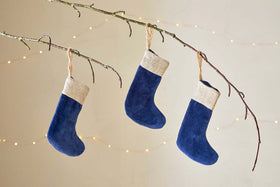 The nkuku Karru Cotton Velvet Mini Stockings - Indigo (Set of 3) hang from a bare branch by twine loops, each with a beige cuff, set against a neutral backdrop with soft string lights for a festive touch.