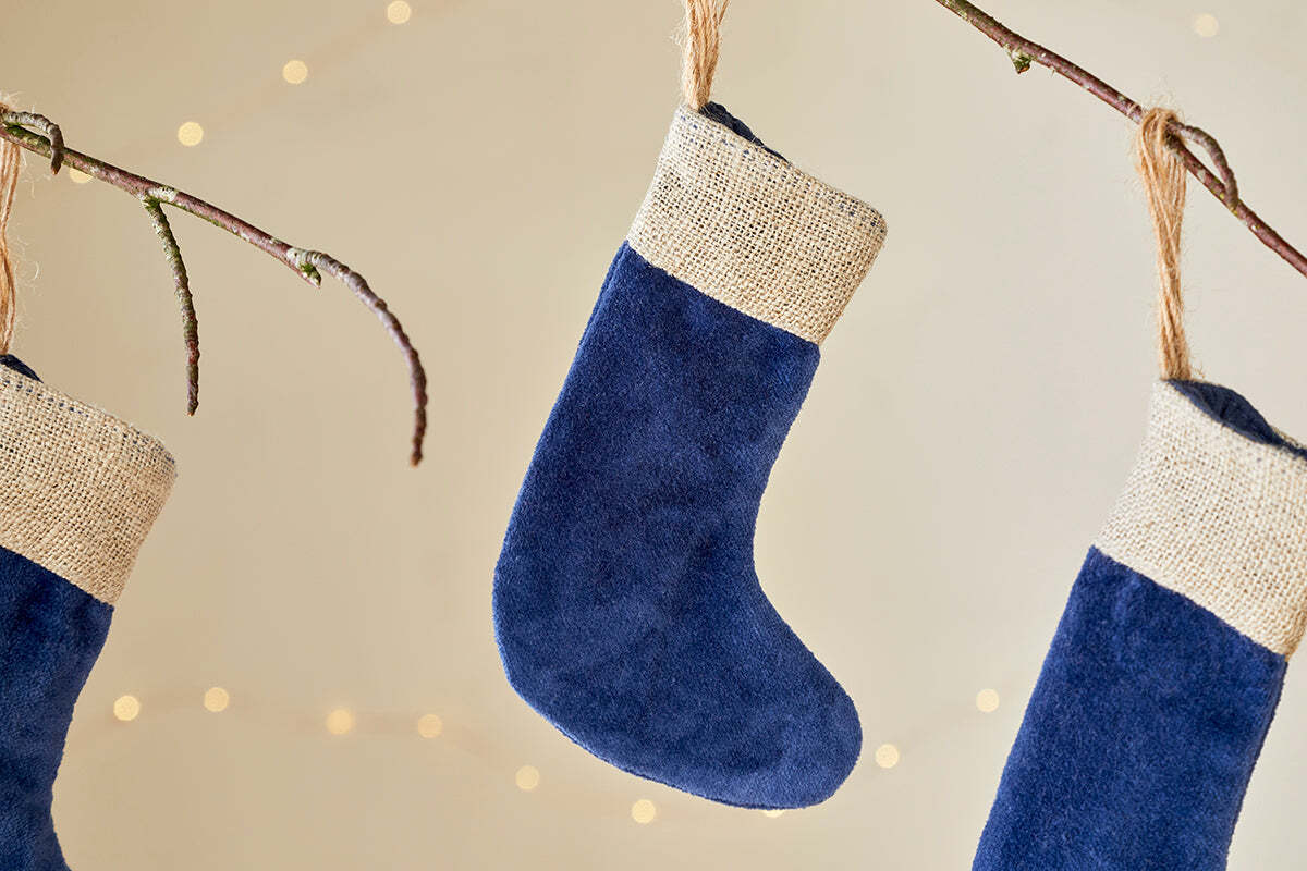 Three Karru Cotton Velvet Mini Stockings in indigo by nkuku (set of 3) hang from bare branches with burlap cuffs and soft string lights, creating a cozy, festive scene against a light background.