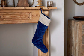 The nkuku Karru Cotton Velvet Stocking in Indigo, featuring a beige cuff, hangs from a rustic wooden mantel surrounded by festive décor like a small tree and candle holders, adding charm to the cozy room.