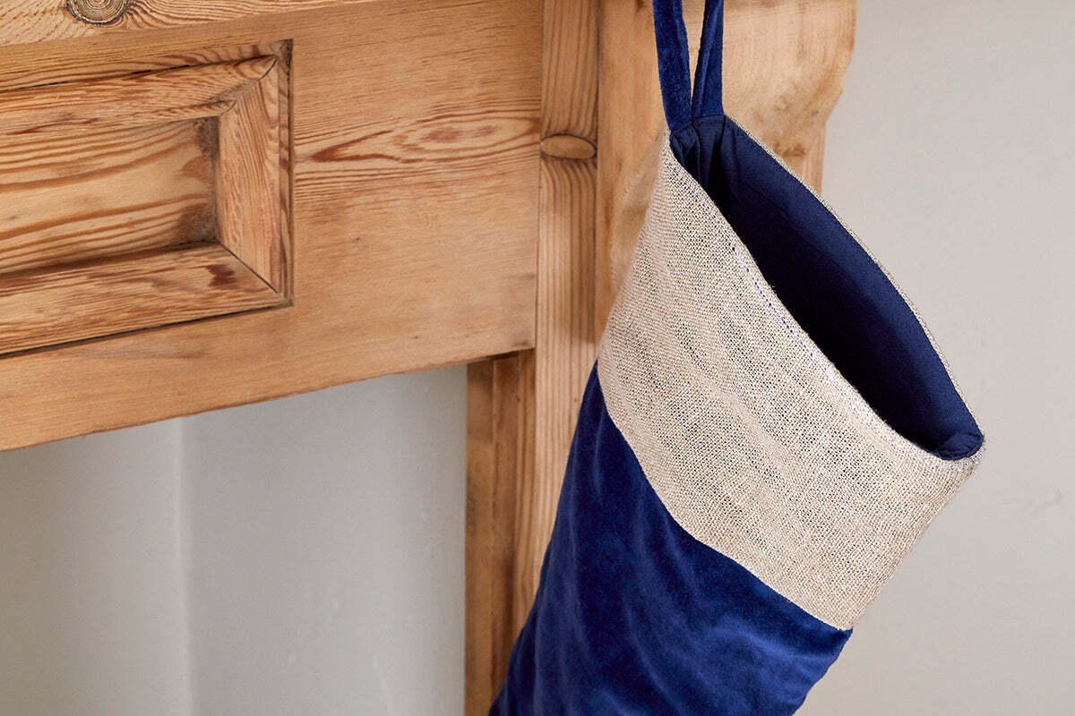 An nkuku Karru Cotton Velvet Stocking in Indigo with a beige woven cuff hangs from a rustic wooden mantel, its natural grain and knots adding charm to the festive scene against a light-colored wall.