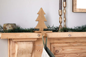 A wooden mantelpiece features green garland, a gold metal tree figurine, two gold candlesticks with brown candles, and a rustic gold candle holder. A stocking hangs below on nkuku's Kotta Stocking Tree Holder in antique brass.