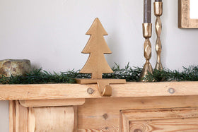 The Kotta Stocking Tree Holder in Antique Brass by nkuku sits on a wooden mantel adorned with green faux pine garland. Two gold candleholders and a small round object are partially seen on either side against a plain white wall.