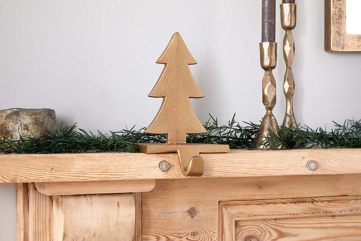 The Kotta Stocking Tree Holder in Antique Brass by nkuku sits on a wooden mantel adorned with green faux pine garland. Two gold candleholders and a small round object are partially seen on either side against a plain white wall.