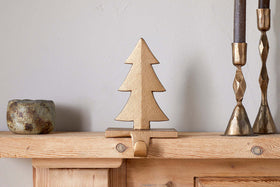 The nkuku Kotta Stocking Tree Holder in antique brass adds charm to a wooden mantel, flanked by two brass candlesticks with dark candles and a rustic ceramic cup, standing out against a plain light-colored wall.
