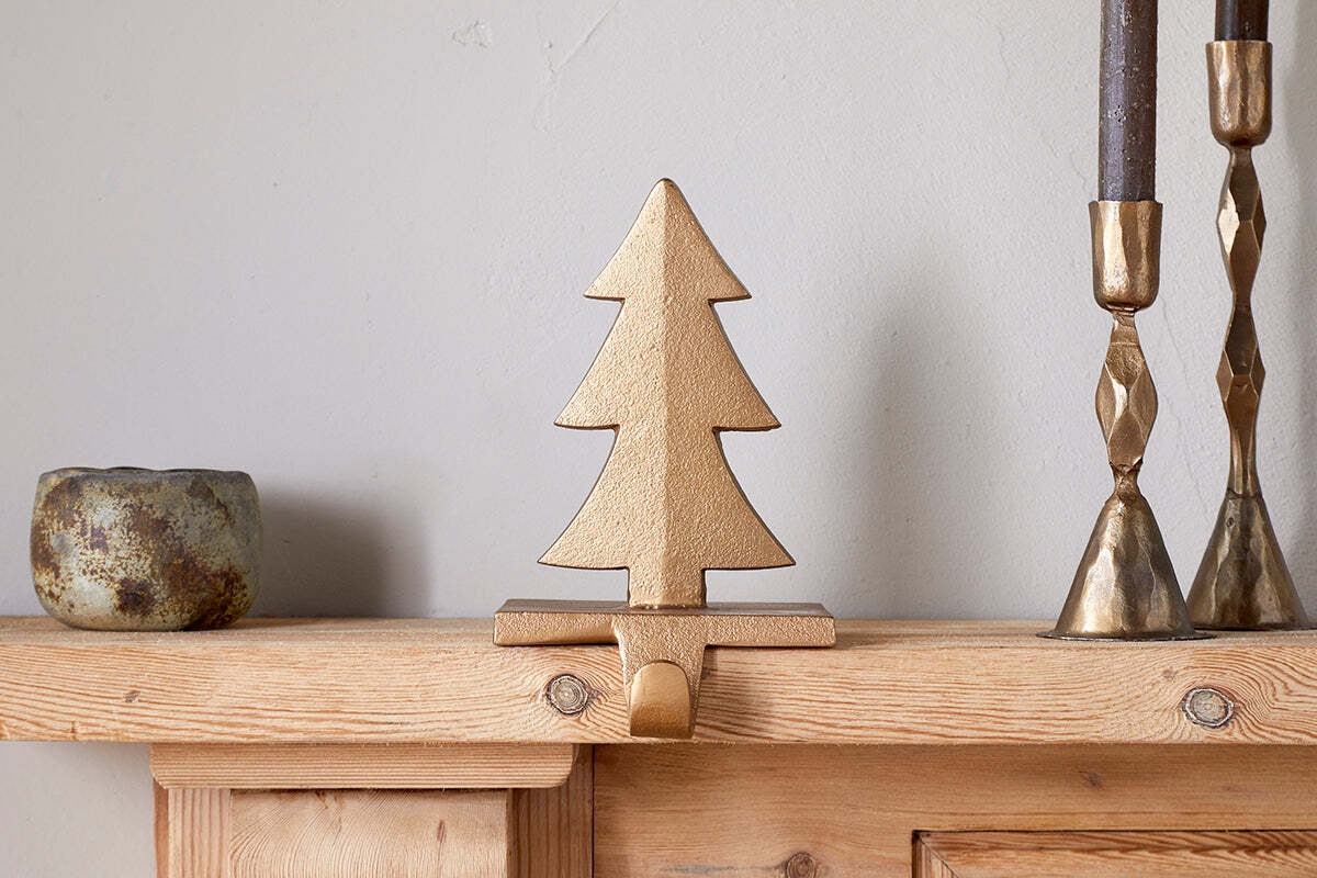 The nkuku Kotta Stocking Tree Holder in antique brass adds charm to a wooden mantel, flanked by two brass candlesticks with dark candles and a rustic ceramic cup, standing out against a plain light-colored wall.