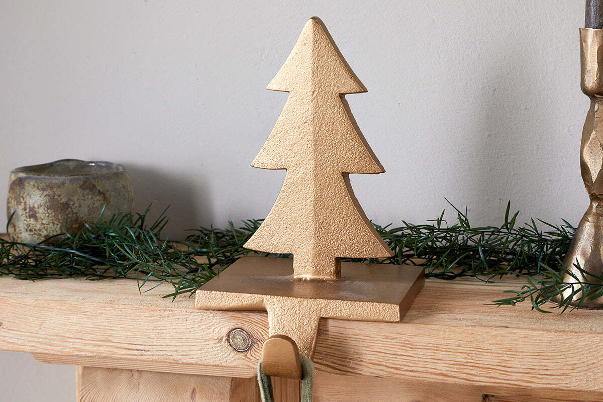 The Kotta Stocking Tree Holder in antique brass by nkuku sits on a wooden mantel with green garland. The brass hook hangs over the edge for stockings, and a small candleholder and candlestick are partially visible nearby.