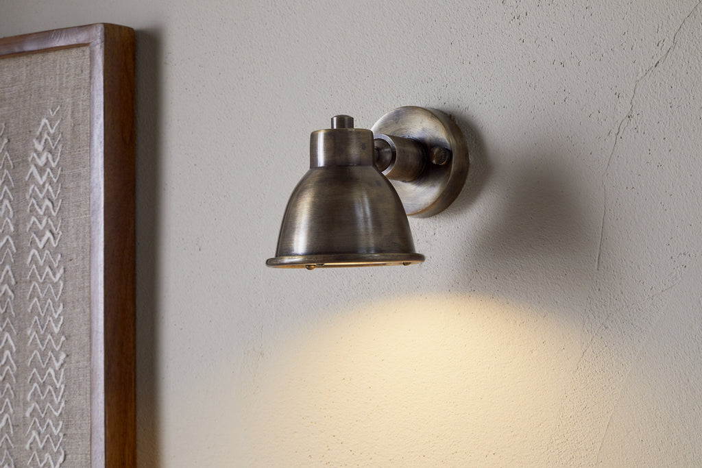 The Kasai Brass Spotlight by nkuku, featuring an antique brass dome shade inspired by industrial design, is mounted on a textured beige wall and casts a warm downward glow. A portion of framed artwork appears at the images left edge.