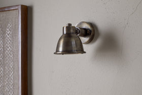 The nkuku Kasai Brass Spotlight in Antique Brass is mounted on a light-colored, textured wall, with part of a framed fabric artwork visible on the left.