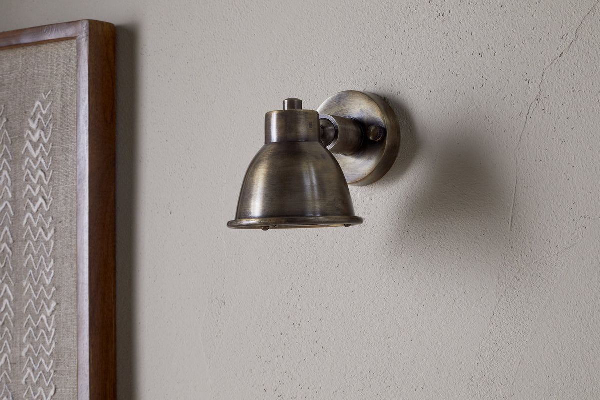 The nkuku Kasai Brass Spotlight in Antique Brass is mounted on a light-colored, textured wall, with part of a framed fabric artwork visible on the left.