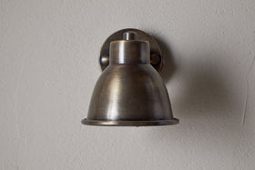 The Kasai Brass Spotlight by nkuku features an antique brass finish and dome-shaped shade, mounted on a light gray textured wall. This adjustable fixture brings vintage industrial style and character to any space.