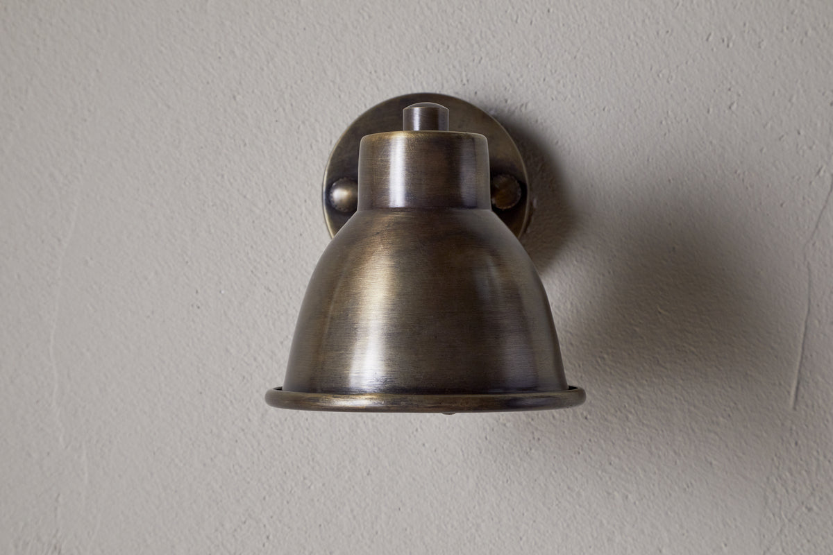 The Kasai Brass Spotlight by nkuku features an antique brass finish and dome-shaped shade, mounted on a light gray textured wall. This adjustable fixture brings vintage industrial style and character to any space.