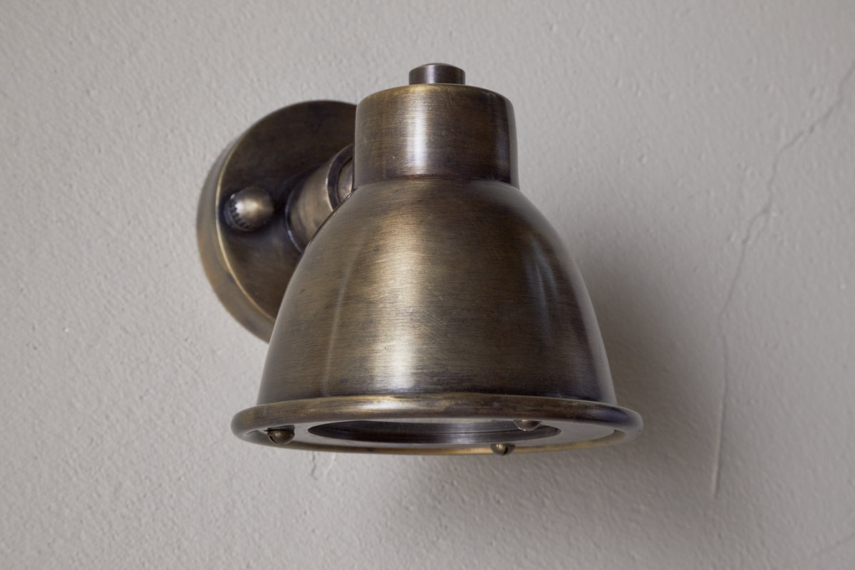 The Kasai Brass Spotlight by nkuku features a dome-shaped, industrial-style antique brass shade on a round wall plate, mounted to a plain light-colored wall for a rustic vintage look.
