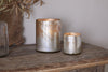 Two metallic Kunjari Tealight Holders in rustic gold from nkuku—one large, one small—rest on a wooden table beside a glass vase of dried leaves, bringing antique charm to the space against a light wall and wooden accent.
