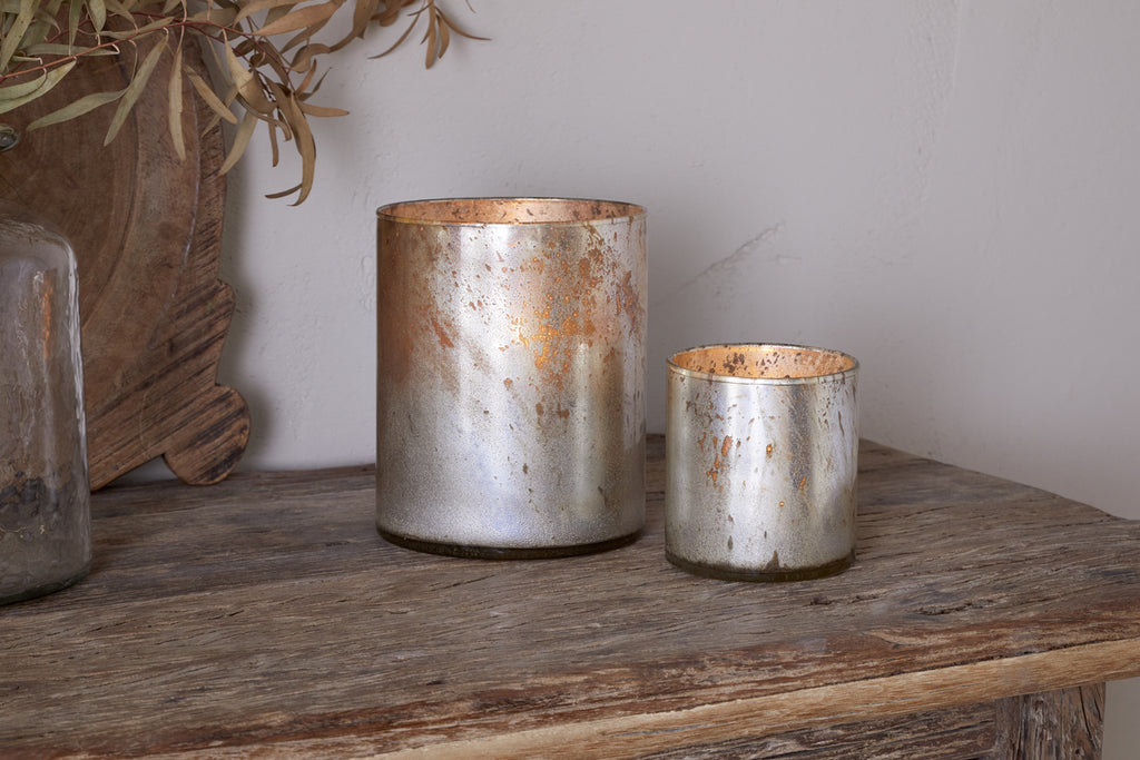 Two metallic Kunjari Tealight Holders in rustic gold from nkuku—one large, one small—rest on a wooden table beside a glass vase of dried leaves, bringing antique charm to the space against a light wall and wooden accent.
