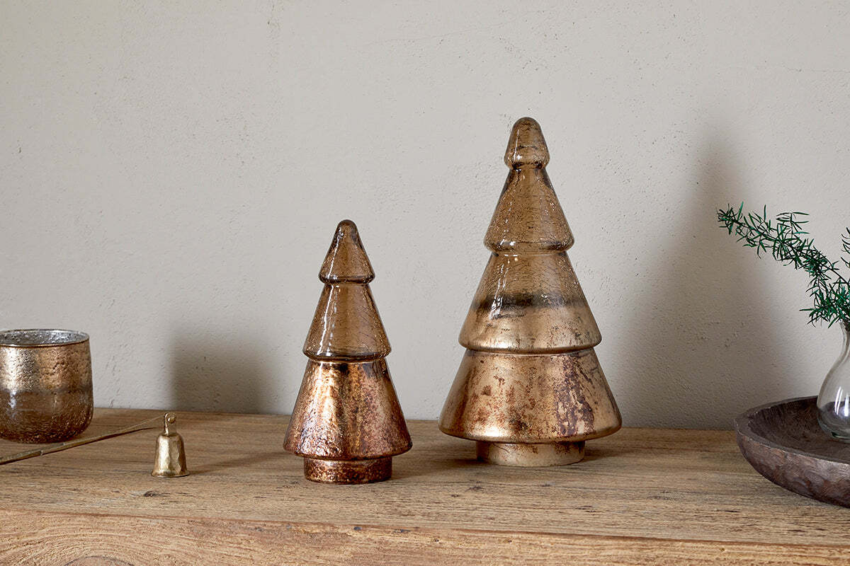Two nkuku Konara Glass Trees in gold crackle add a festive touch on a wooden surface. The larger tree sits on the right beside a gold bell, glass candle holder, and vase with greenery against a plain wall.