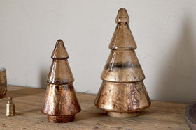 Two metallic, cone-shaped Konara Glass Trees in gold crackle by nkuku sit on a wooden surface against a beige wall. Each gold tree adds a striking, festive accent to your holiday decor.