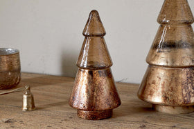 The Konara Glass Tree - Gold Crackle by nkuku sits on a wooden surface beside a larger gold tree, with a bell-shaped ornament and silver tumbler nearby, creating festive, rustic decor.