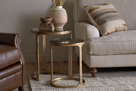 The Khidki Cast Metal & Glass Nesting Side Tables by nkuku in antique brass are placed beside a beige sofa with a patterned pillow, topped with a vase of dried flowers and wooden decor. A leather armchair and textured rug complete the look.