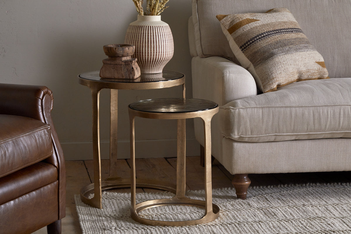 The Khidki Cast Metal & Glass Nesting Side Tables by nkuku in antique brass are placed beside a beige sofa with a patterned pillow, topped with a vase of dried flowers and wooden decor. A leather armchair and textured rug complete the look.