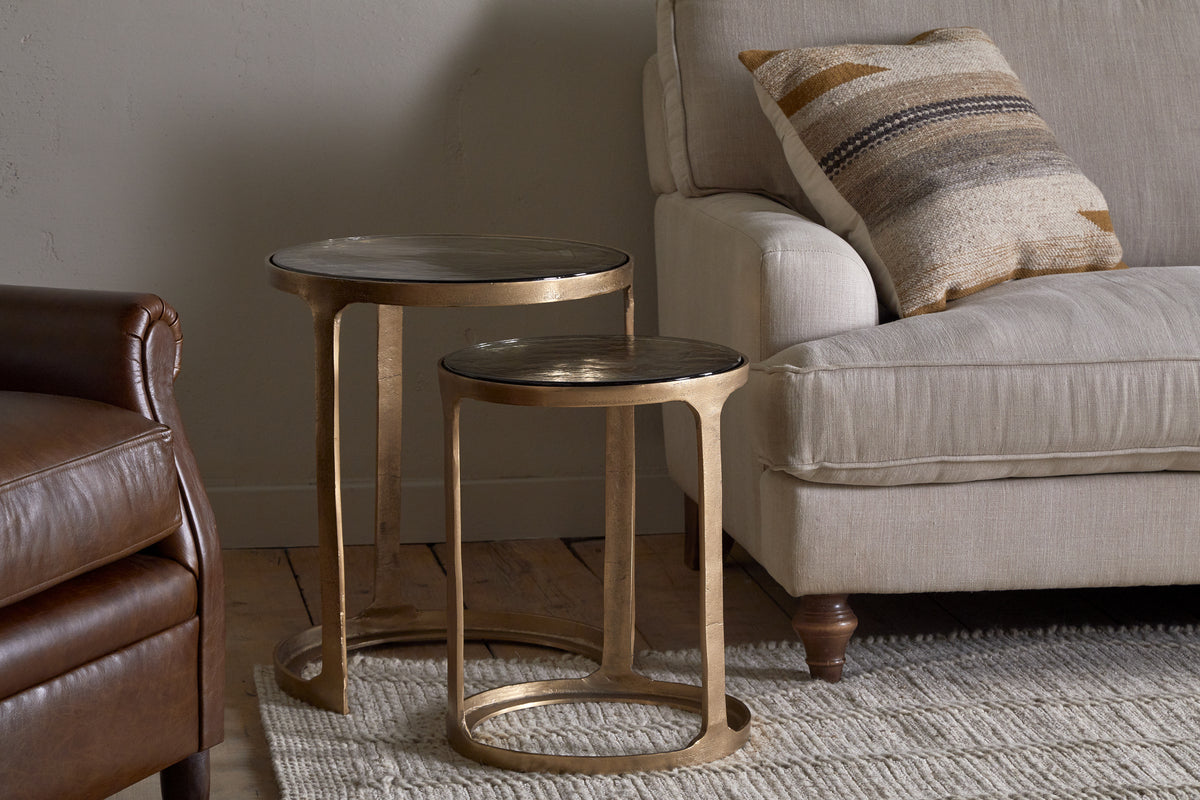 The Khidki Cast Metal & Glass Nesting Side Tables by nkuku, featuring antique brass frames, are placed beside a beige sofa with a patterned cushion, next to a leather armchair on a light woven rug.