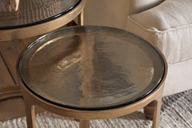 The Khidki Cast Metal & Glass Nesting Side Tables by nkuku, featuring antique brass frames and glass tops, sit beside a beige upholstered chair on a textured light rug.