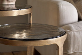 Close-up of the Khidki Cast Metal & Glass Nesting Side Tables (set of 2) in antique brass by nkuku, featuring a shiny hammered finish, shown beside a beige upholstered sofa in a cozy living room.