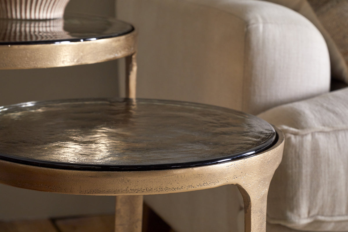 Close-up of the Khidki Cast Metal & Glass Nesting Side Tables (set of 2) in antique brass by nkuku, featuring a shiny hammered finish, shown beside a beige upholstered sofa in a cozy living room.