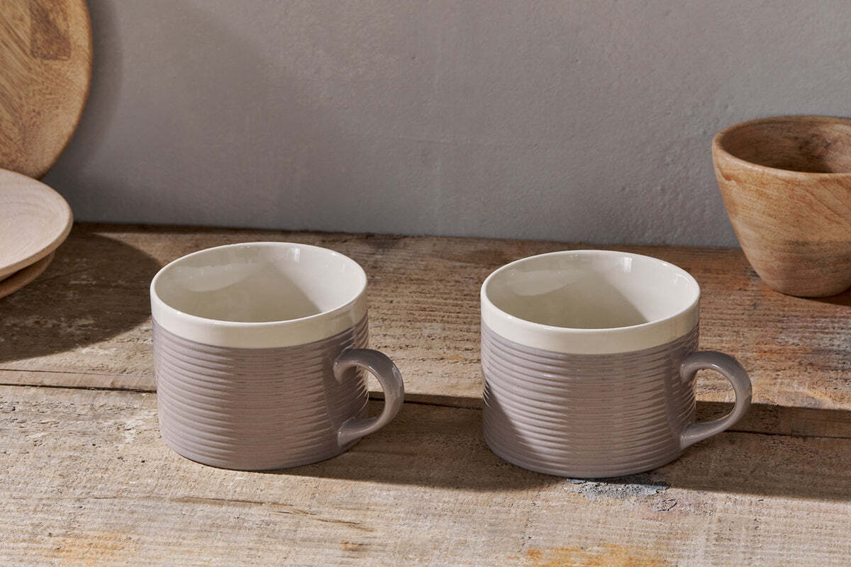 The nkuku Kai Mugs - Grey (Set of 2) feature a contemporary ribbed design and white interiors, displayed on a rustic wooden surface with wooden bowls and plates in the background for a cozy, natural kitchen vibe.