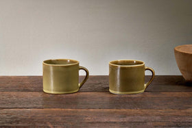 A set of two small green Kalini mugs by nkuku with handles are placed side by side on a rustic wood surface, with a brown ceramic bowl partly visible on the right and a plain, light-colored wall in the background.