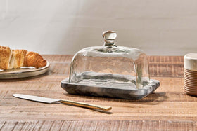 The nkuku Kallso Marble Butter Dish rests on a wooden table, its recycled glass lid with a marble base. Beside it are a gold-handled butter knife, a plate holding a croissant, and a ridged brown-and-white ceramic cup.