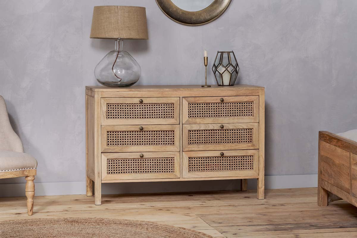 The nkuku Kanz Mango Wood Chest Of Drawers - Natural features six rattan drawers for stylish storage. A glass lamp with burlap shade, brass candlestick, and geometric lantern are on top; a tufted armchair sits left, wooden bed frame right.