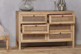 The nkuku Kanz Mango Wood Chest Of Drawers - Natural features cane panels and rattan drawer fronts for stylish storage. Two top drawers are open, revealing more inside. To the left sits a chair with a light fabric seat and carved legs.