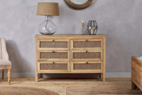 The nkuku Kanz Mango Wood Chest Of Drawers - Natural, featuring six wicker-fronted drawers, stands against a light gray wall. On top are a glass lamp with a tan shade, gold candleholder, and decorative lantern for stylish bedroom storage.
