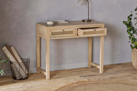 The nkuku Kanz Mango Wood Writing Desk - Natural stands against a gray wall, topped with a lamp, cup, and book. Rolled papers in a wire basket, a braided rug, and green plants accent the floor—ideal home office furniture.