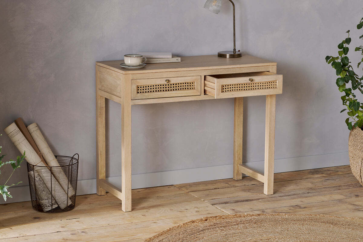 The nkuku Kanz Mango Wood Writing Desk - Natural stands against a gray wall, topped with a lamp, cup, and book. Rolled papers in a wire basket, a braided rug, and green plants accent the floor—ideal home office furniture.