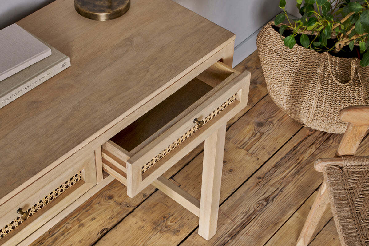 The Kanz Mango Wood Writing Desk by nkuku features an open cane panel drawer and lattice trim, sitting on a rustic wooden floor. A woven basket plant, woven-seat chair, closed book, and glass lamp complete this inviting setup.