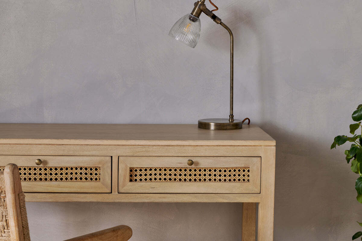 The Kanz Mango Wood Writing Desk by nkuku, featuring cane panel drawers, is set against a light gray wall. A brass lamp with a glass shade rests on the desk, with part of a wooden chair and a green plant visible in the foreground.