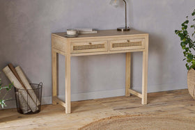 The nkuku Kanz Mango Wood Writing Desk - Natural stands against a gray wall, topped with a book, cup, and lamp. A wire basket with rolled papers sits to the left and green plants to the right. A round woven rug lies on the floor.