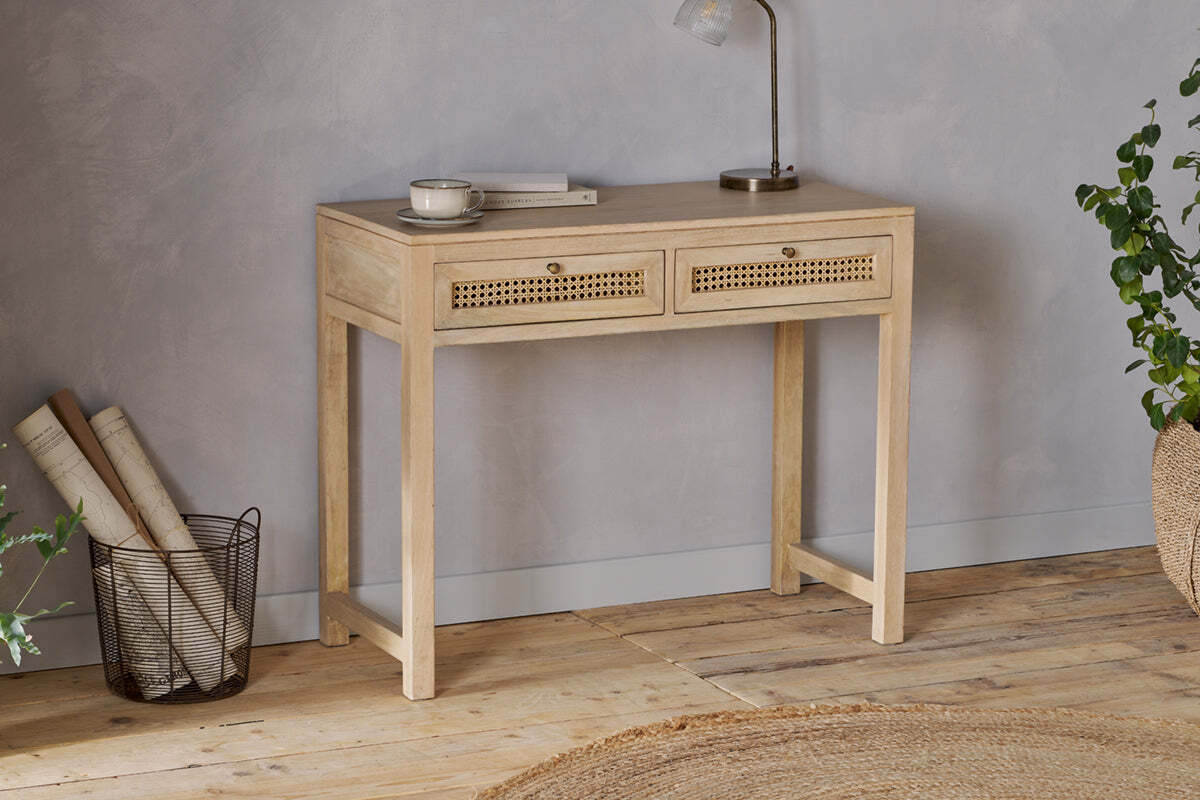 The nkuku Kanz Mango Wood Writing Desk - Natural stands against a gray wall, topped with a book, cup, and lamp. A wire basket with rolled papers sits to the left and green plants to the right. A round woven rug lies on the floor.