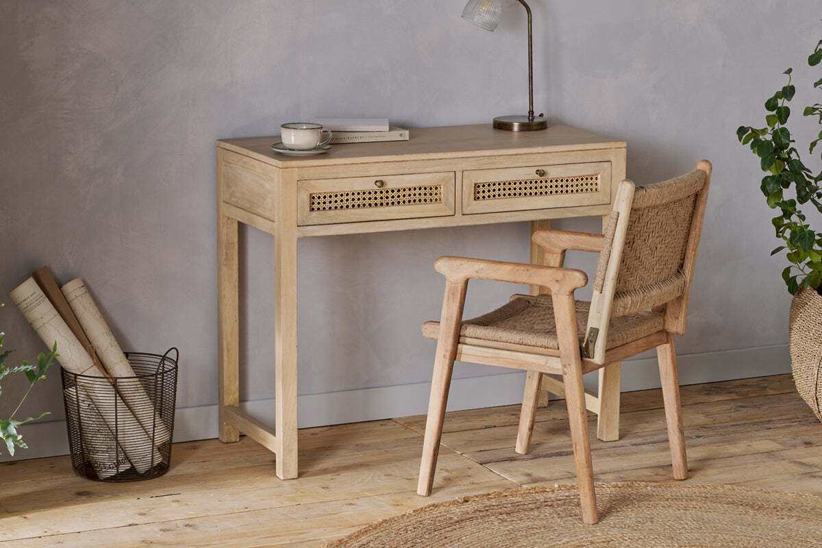 The nkuku Kanz Mango Wood Writing Desk - Natural stands against a gray wall, paired with a matching chair. Rolled papers, a plant, and a round rug on the wooden floor complete this cozy home office setup.