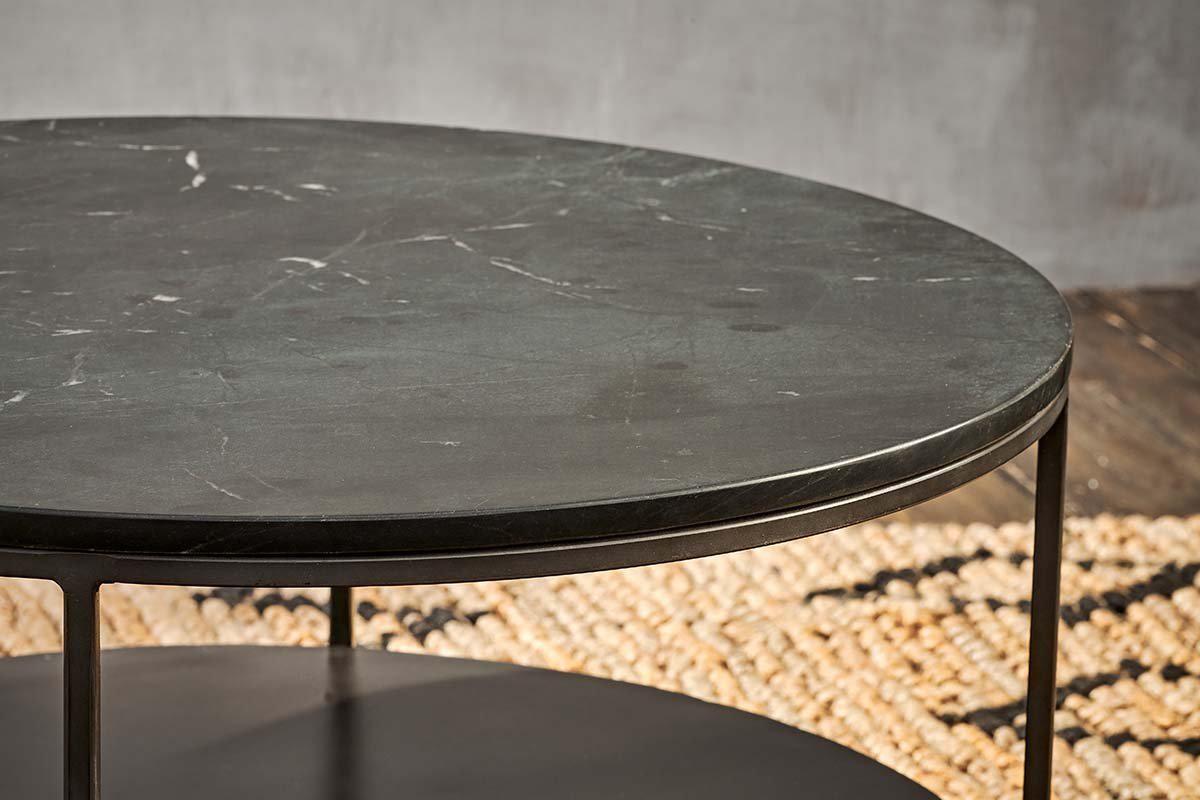 A close-up of the Karoko Marble Coffee Table by nkuku, featuring a round black marble top with white veining and slim iron legs, placed on a woven natural fiber rug over dark wood floors against a textured gray wall.