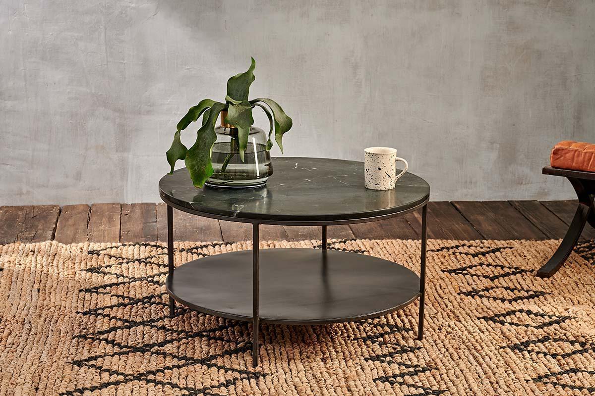 The Karoko Marble Coffee Table by nkuku, featuring a round black marble top and iron framework, holds a glass vase with green foliage and a white speckled mug. It sits on a geometric woven rug near an orange-cushioned chair and gray wall.