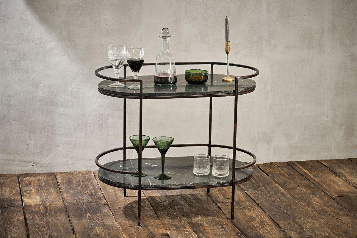 The Karoko Marble Drinks Table by nkuku features two oval black marble shelves and a distressed metal frame, perfect for displaying glassware, a decanter of red wine, a green bowl, and a gold candlestick with a grey candle.