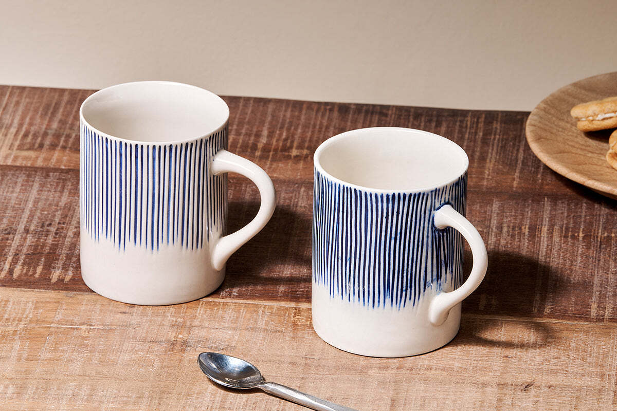 Two white nkuku Karuma Ceramic Mugs - Large (Set of 2) with blue vertical stripes sit on a wooden table, accompanied by a metal teaspoon. A wooden plate with cookies appears partially in the background on the right.