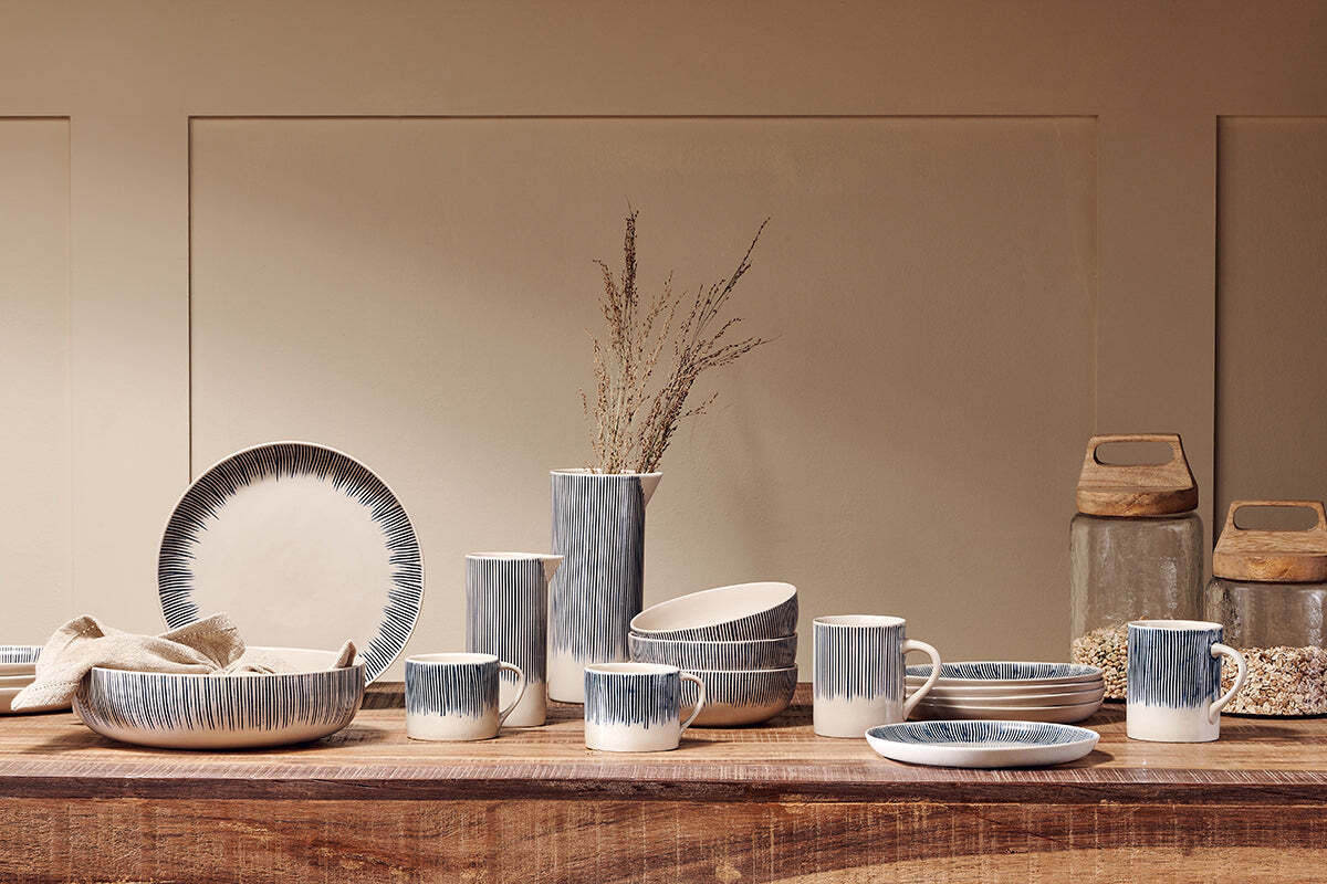 A wooden table showcases nkuku’s Karuma Ceramic Mug - Large (Set of 2) alongside handmade bowls, dishes, and a vase with dried grass. The blue-gray and white gradients create a modern rustic style, with two jars of grains nearby against a beige wall.